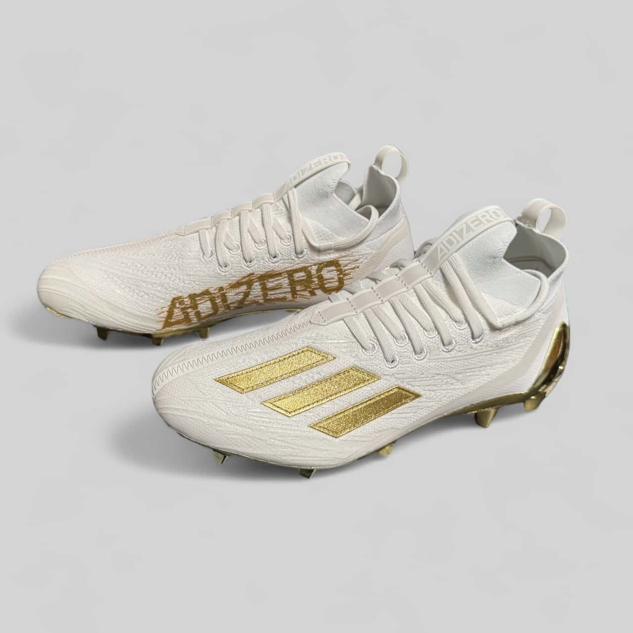 Zapato Adidas Adizero Primeknit blanco y dorado, diseñado para béisbol con tejido flexible y suela metálica para tracción superior.